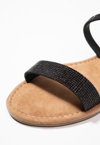 Mustat helmillä varustetut sandaalit, joissa on pehmeä vaaleanruskea mokkanahkainen pohja; tasapohjalla ja huolellisella ompelulla, joka korostaa käsityötä ja yksityiskohtia.