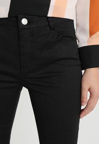 Gros plan sur une personne portant un pantalon noir pailleté avec un bouton blanc et un haut à manches longues à rayures verticales beige, orange et noir.