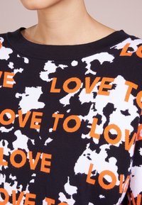 Pull-over à motifs noir et blanc avec le texte orange "LOVE TO LOVE" en caractères gras. Tissu doux avec un col décontracté, présentant une surface texturée.