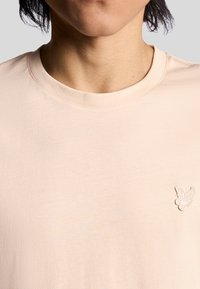 Lichtbeige katoenen t-shirt met een ronde halslijn. Heeft een kleine witte vlinder-geborduurde op de linkerborst. Zachte textuur.