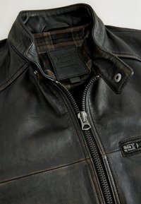 Veste en cuir noire avec une fermeture éclair métallique, un col à pression et une doublure en tissu à carreaux visible à l’intérieur du col.