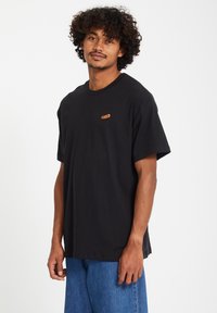 Zwart katoenen t-shirt met een ronde hals, losse pasvorm en een klein oranje logo op de borst. De stof lijkt zacht en heeft een gladde textuur.