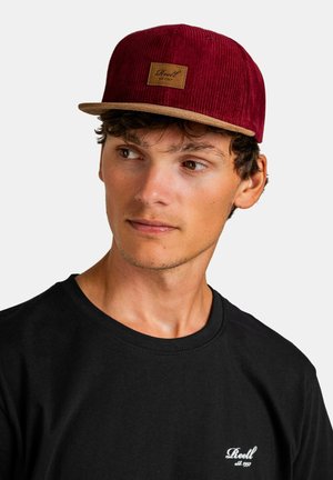 Bordeauxfarbenes Cord-Hut mit einem hellbraunen Lederschild, flachemschirm und strukturiertem Stoff. Kombiniert mit einem schlichten schwarzen T-Shirt.