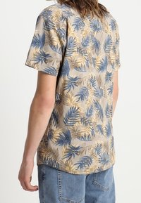 Short-sleeve shirt featuring a beige base with a blue and gold tropical leaf pattern. Lightweight fabric with a relaxed fit and soft texture.