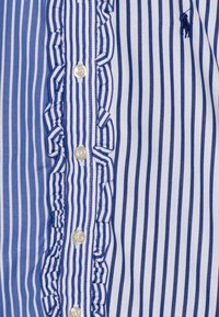 Blå och vitrandig skjorta med en volangklädd placket, vita knappar och en liten broderad logo på tyget.