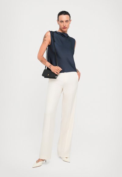 Sleeveless navy top with cowl neckline, paired with white tailored pants. Accessorized with a small black crossbody bag and heeled shoes.