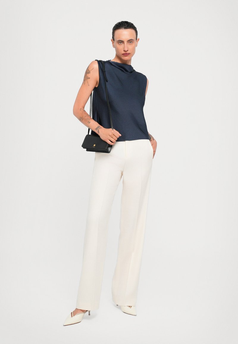 Ermeløs navy top med draperet hals, kombineret med hvide skræddersyede bukser. Accessorieset med en lille sort crossbody taske og hæle sko.