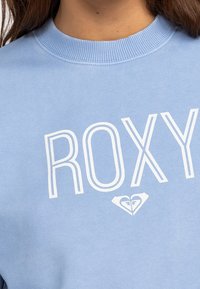 Felpa azzurra chiaro con grande scritta bianca "ROXY" e una piccola grafica a forma di cuore sotto il testo, con scollatura a coste.