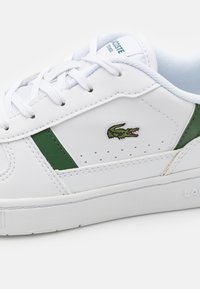 Zapatilla blanca hecha de cuero texturizado con acentos verdes. Cuenta con un logo de cocodrilo bordado en verde, cordones redondos y paneles laterales perforados.
