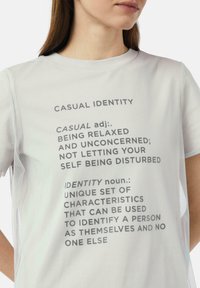 Jasnoszara koszulka z okrągłym dekoltem i krótkimi rękawami, z tekstowymi definicjami słów "casual" i "identity" w pogrubionym czarnym druku.