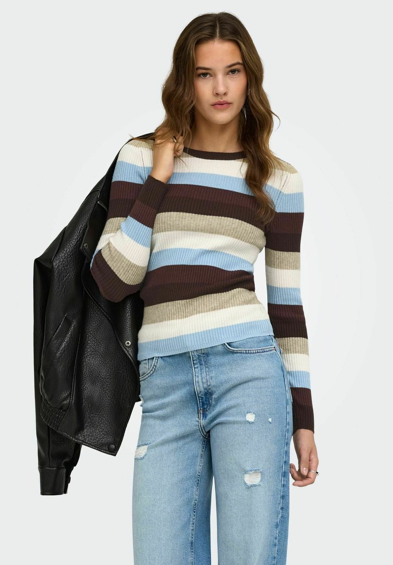 Jeune femme portant un pull à rayures et un jean déchiré tient une veste en cuir noire sur son épaule devant un fond uni.