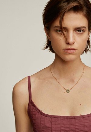 Collar de cadena dorada con una piedra verde circular, llevado con una blusa burdeos ajustada que muestra detalles de costura vertical.