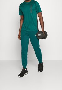 Tyrkysový sportovní top s černými akcenty, tyrkysové tepláky a černé tenisky, držící činky, na tričku a kalhotách je logo Puma.