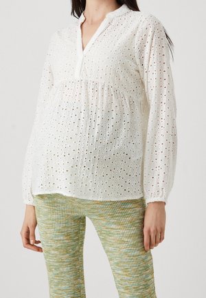 Kobieta ubrana w białą bluzkę z ażurowego materiału z długim rękawem i dekoltem w kształcie litery V oraz zielone spodnie w wzory, stojąca na jednolitym tle.