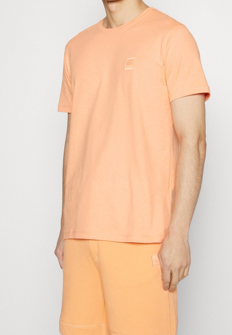 Meruňková bavlněná tričko s krátkými rukávy, kulatým výstřihem a malým čtvercovým logem na prsou. Hladká textura, volný střih.