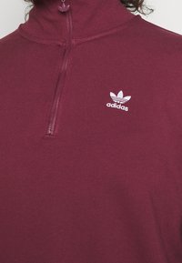 Primer plano de una persona que lleva una parte superior con cremallera color burdeos y el logo blanco de Adidas en el lado izquierdo del pecho, con el cabello rizado parcialmente visible.