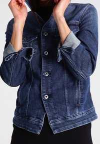 Person som justerar huvan på en blå denimjacka med uppvikta ärmar och metallknappar, iklädd svarta byxor.