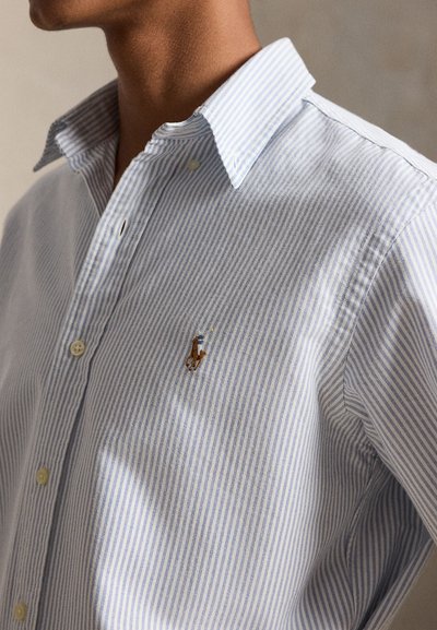 Nærbilde av en person som har på seg en lyseblå og hvitstripet skjorte med knapper og en liten brodert polospiller-logo på brystet.