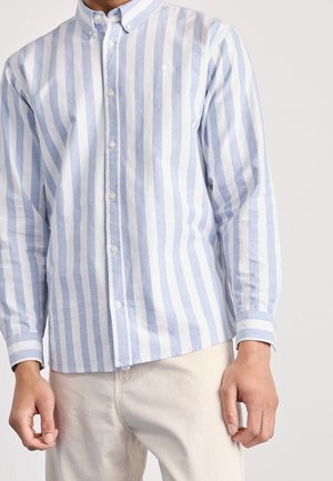 Mann trägt hellblau-weiß gestreiftes Hemd mit langen Ärmeln und beigen Hosen, steht vor einfarbigem Hintergrund.