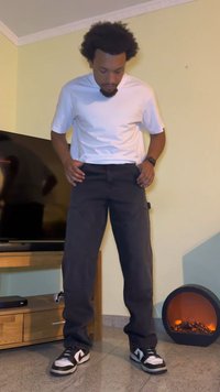 Black cargo pants with pockets, paired with a plain white t-shirt. Black and white low-top sneakers. Background includes a TV and decorative heater.