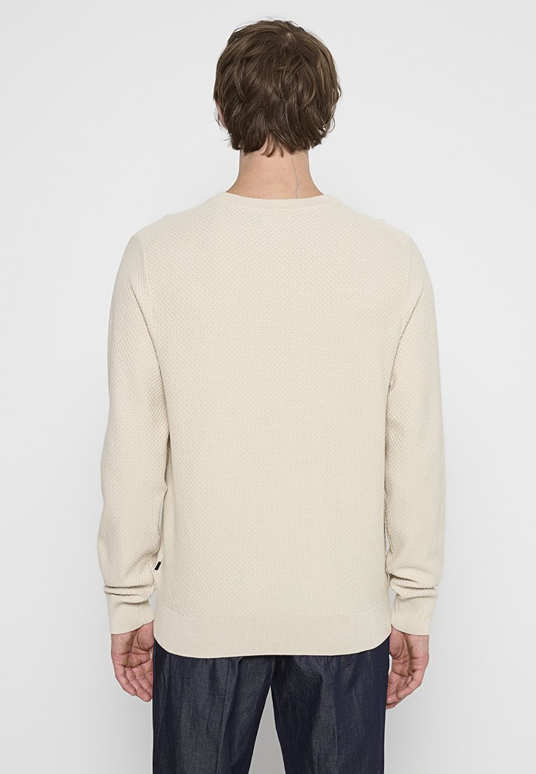 Man med kort brunt hår bär en texturerad krämfärgad stickad tröja och mörka byxor, står vänd bortåt mot en helt vit bakgrund.