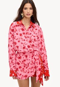 Blouse rose à motif floral avec accents rouges et manches longues, dotée d'une poche et d'un détail noué à la taille. Tissu lisse avec finition brillante.