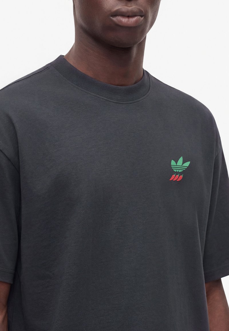 Man wearing a dark gray T-shirt with a small green and red logo on the left chest showing a stylized leaf and three chili peppers.