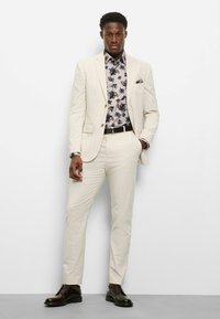Light beige suit with a slim fit; patterned floral shirt; dark belt; sleek brown shoes; accessories include a pocket square.