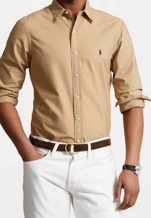 Homme portant une chemise beige boutonnée avec manches retroussées, un pantalon blanc, une ceinture marron et une montre avec un bracelet multicolore au poignet gauche.