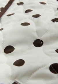 Prešívaná látka s bielym pozadím a veľkými okrúhlymi hnedými bodkami; hladký povrch so subtílnym leskom a kontrastným prešívaním.