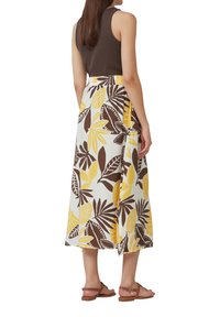 Bruine mouwloze top gecombineerd met een middellange rok met een witte achtergrond en gele en bruine bladenpatronen. Lichtbruine sandalen.