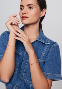 Koszula dżinsowa z krótkim rękawem, noszona przez modela rąk. Dwa bransoletki: jedna z kolorowymi koralikami, a druga z małymi ciemnymi koralikami.