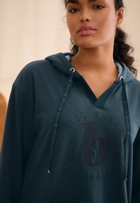 Donkergroene hoodie met een V-hals, voorzien van een getextureerd logo in het midden en contrasterende koordjes met metalen uiteinden. Zacht materiaal.