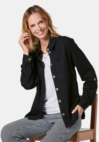 Schwarze Hemdjacke aus Stoff mit durchgängiger Knopfleiste, silbernen Beschlägen, langen Ärmeln und einer Brusttasche. Weißes Oberteil und karierte Hose.