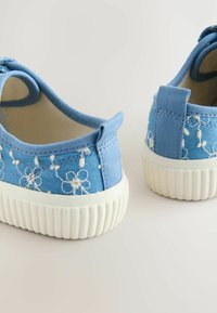 Baskets en toile bleue avec broderie florale blanche, panneaux latéraux élastiques et semelle en caoutchouc blanche avec un design texturé en bas.