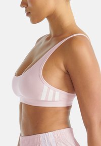 Kobieta w jasnoróżowym staniku sportowym z białymi paskami i dopasowanych szortach sportowych, pokazana z boku na jednolitym tle.