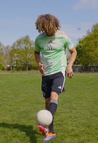 Groen en wit voetbalshirt met 'Italia' logo, zwarte shorts en meerkleurige voetbalschoenen, terwijl hij op gras een witte voetbal dribbelt.