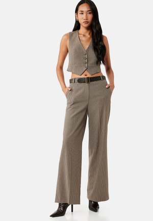 Mujer que lleva un chaleco sin mangas marrón de cuadros y pantalones de pierna ancha a juego con un cinturón negro y tacones negros de punta afilada, de pie con las manos en los bolsillos.