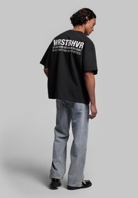 Black oversized cotton t-shirt with bold white text on the back. Paired with light blue straight-leg jeans and black footwear.