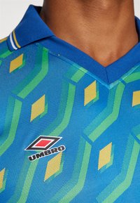 Sinine polo särk, millel on geomeetriline muster rohelises ja kollases. Krae on sinine kollaste ja valgete triipudega. Tikkitud Umbro logo.