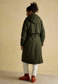 Długi, oliwkowozielony wodoodporny płaszcz z kapturem, z wcięciem w talii i elastycznymi mankietami, zestawiony z białymi spodniami i pomarańczowymi botkami za kostkę.