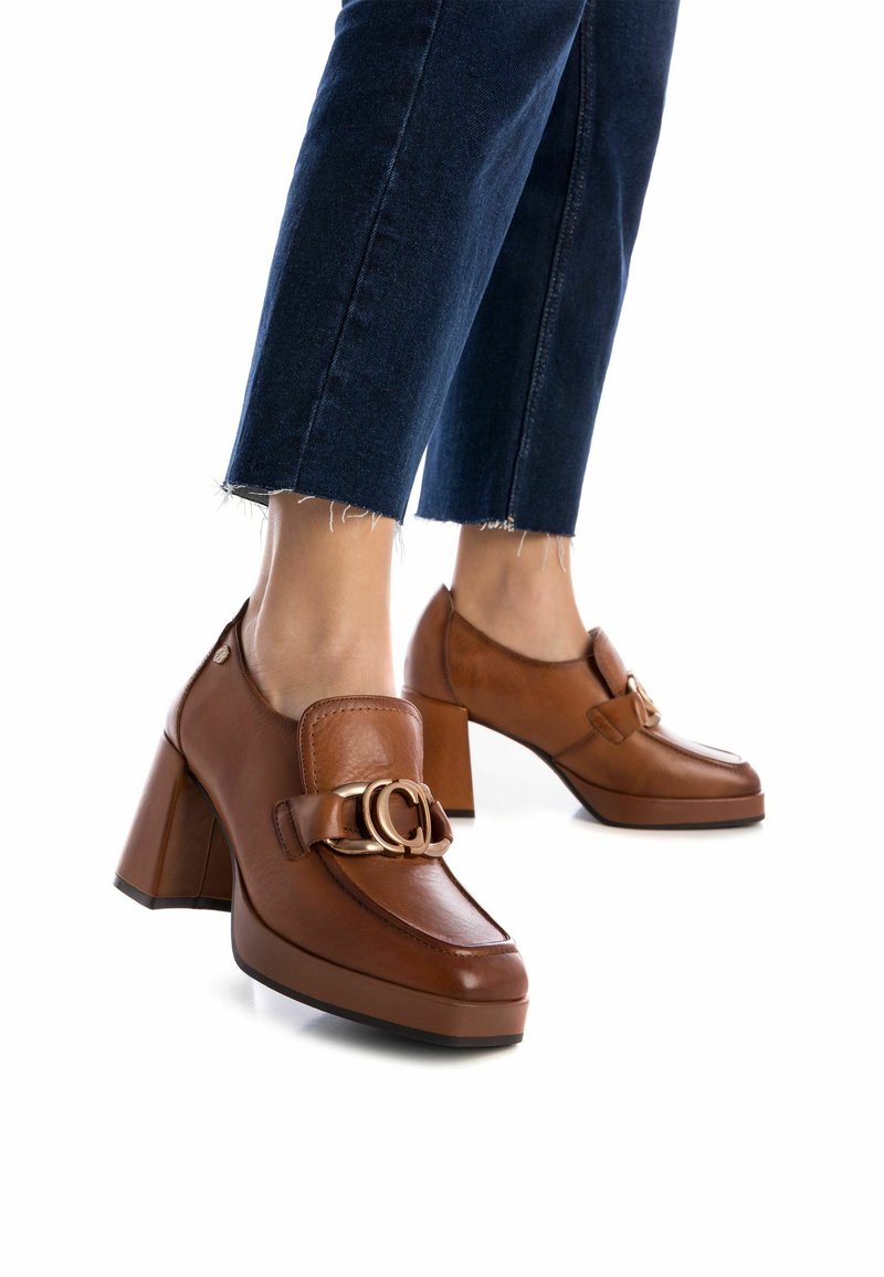 Mocasines de cuero marrón con tacón, punta cuadrada, gran hebilla dorada y tacón ancho. Usados con pantalones de mezclilla oscuros y recortados.