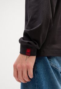 Maglietta nera a maniche lunghe con dettagli ai polsini. Presenta un'etichetta rossa con il logo "HUGO". Mano che afferra jeans di denim con cuciture visibili.