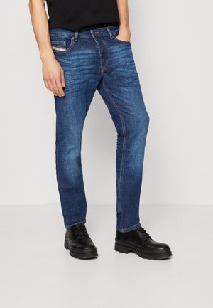 Mężczyzna ubrany w niebieskie dopasowane jeansy, czarną koszulę i czarne sznurowane buty, stojący z jedną ręką w kieszeni na jednolitym tle.