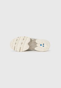 Beige New Balance lenkkarin pohja, jossa on teksturoitua pitoa ja sinisiä vaimennuselementtejä tasaisella valkoisella taustalla.