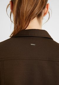 Ruskea takki, jossa on teksturoitu pinta. Varustettu kauluksella ja näkyvillä ompeleilla. Pieni hopeinen "RAW" logo takana, lähellä kaula-aukkoa.