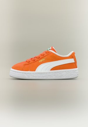 Orange Puma-sneaker med hvid sål og hvid sidestribe, gyldent Puma-logo, fotograferet fra siden på ensfarvet baggrund.