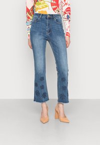 Modré denimové džíny s vysokým pasem, rozšířeným střihem a vyšívanými květinovými vzory na lemu. V kombinaci s nahými lodičkami na vysokém podpatku.