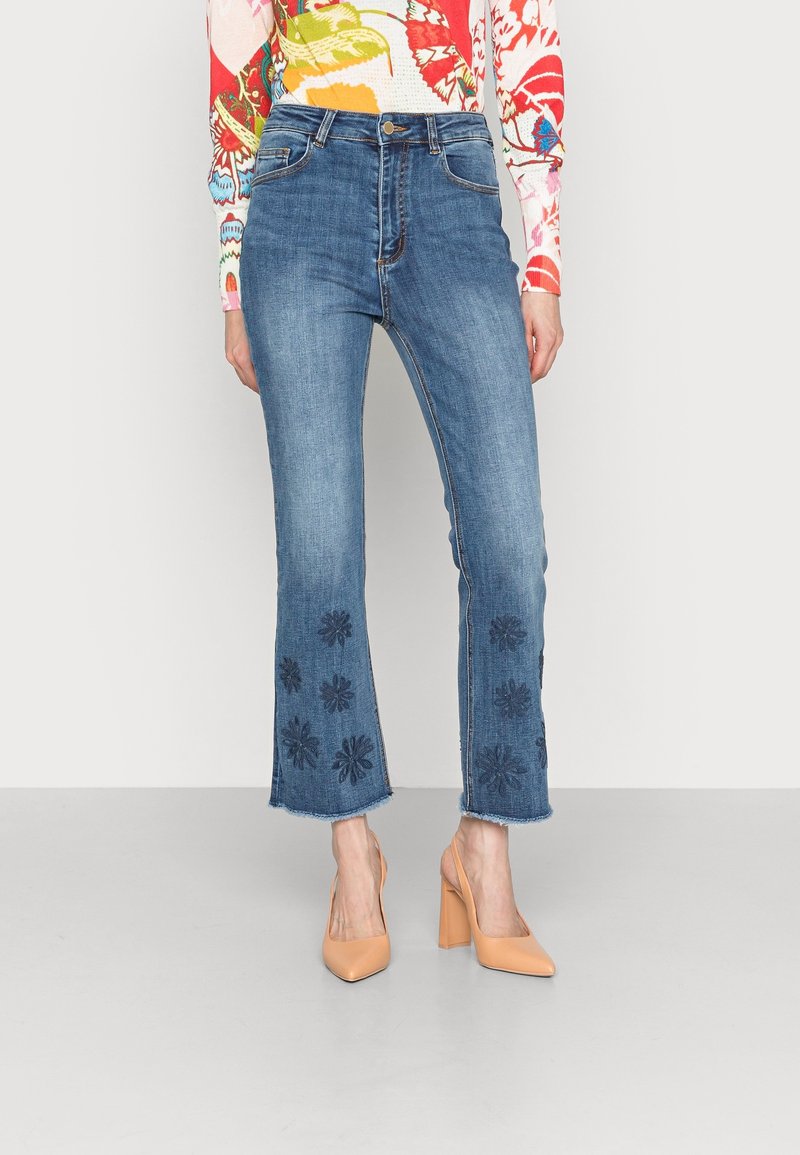 Modré denimové džíny s vysokým pasem, rozšířeným střihem a vyšívanými květinovými vzory na lemu. V kombinaci s nahými lodičkami na vysokém podpatku.