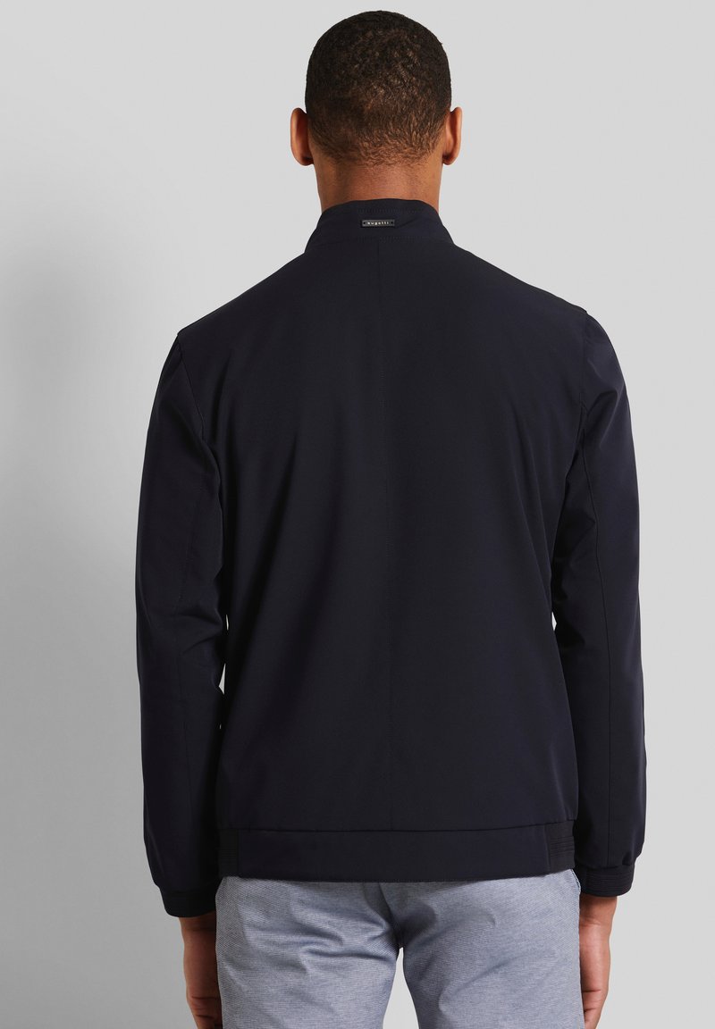Veste légère bleu marine avec un col montant, surface texturée et poignets élastiques, portée sur un pantalon gris. Design simple et moderne.